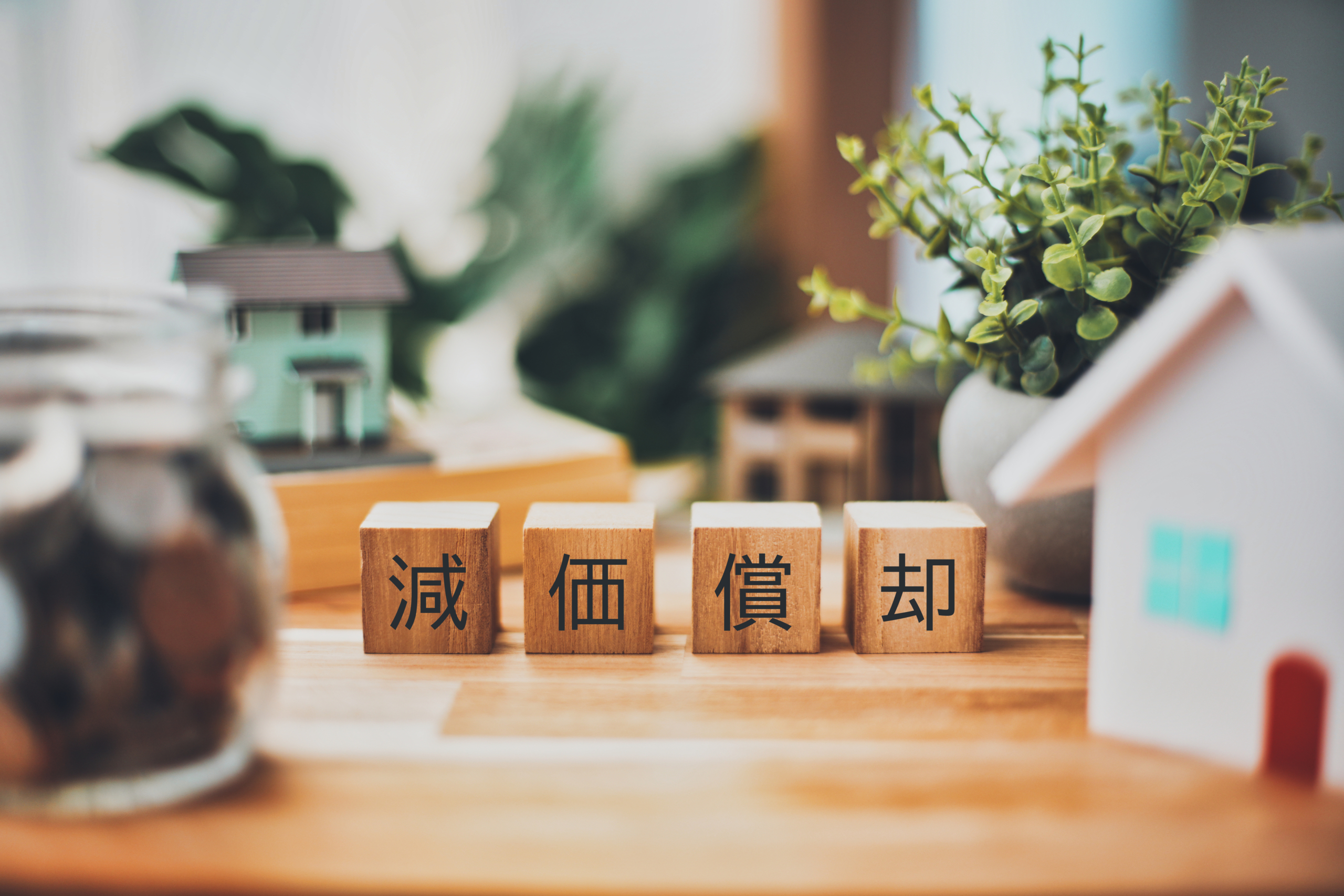 「減価償却」と記された木製のブロックが並び、周囲に住宅のミニチュア模型、観葉植物、硬貨が入った瓶が配置されている。不動産投資や税務を連想させるビジネスイメージ。