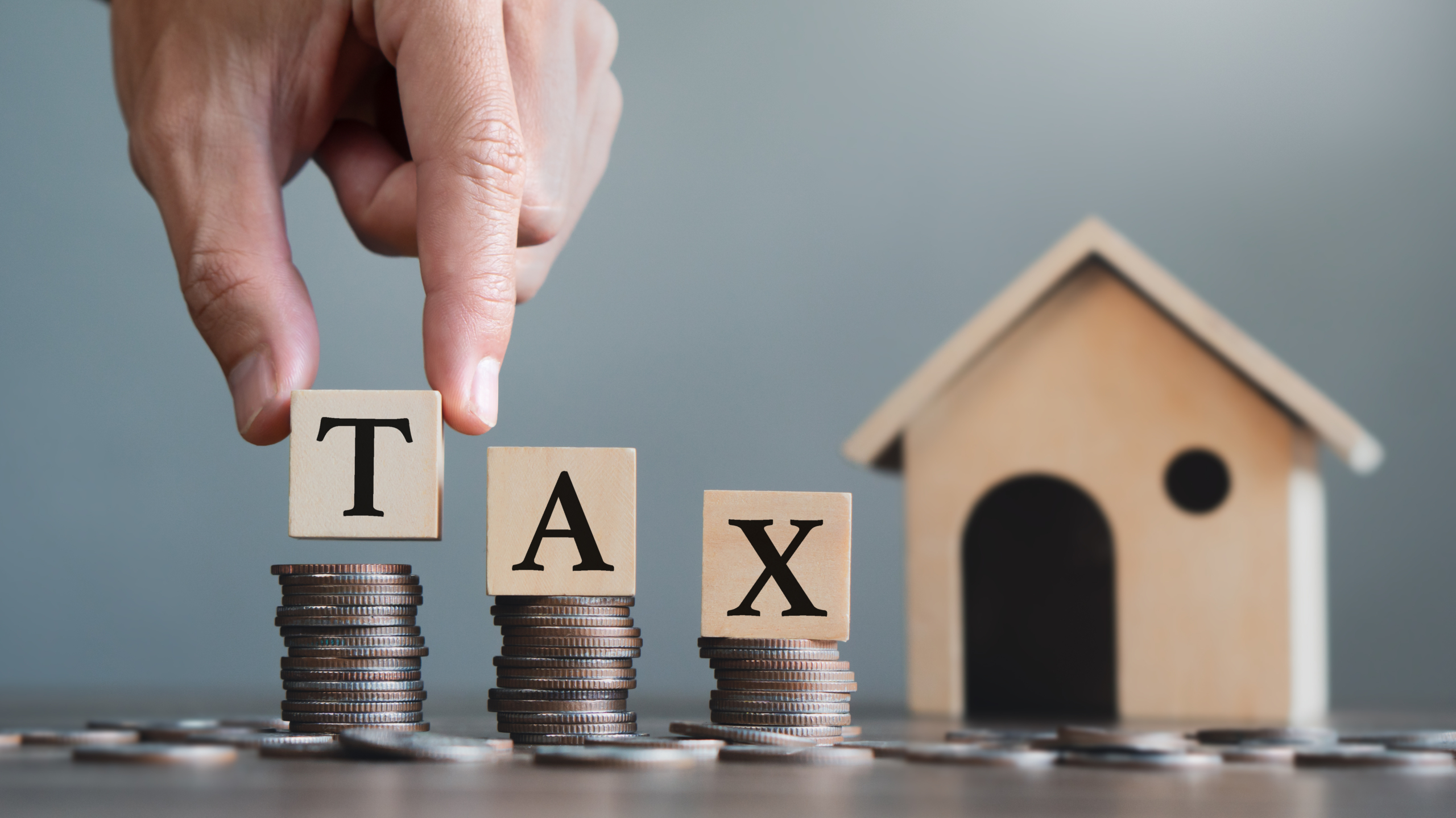 積み上げられたコインの上に『TAX』の木製ブロックを置く手と、その隣に並ぶ住宅のミニチュア