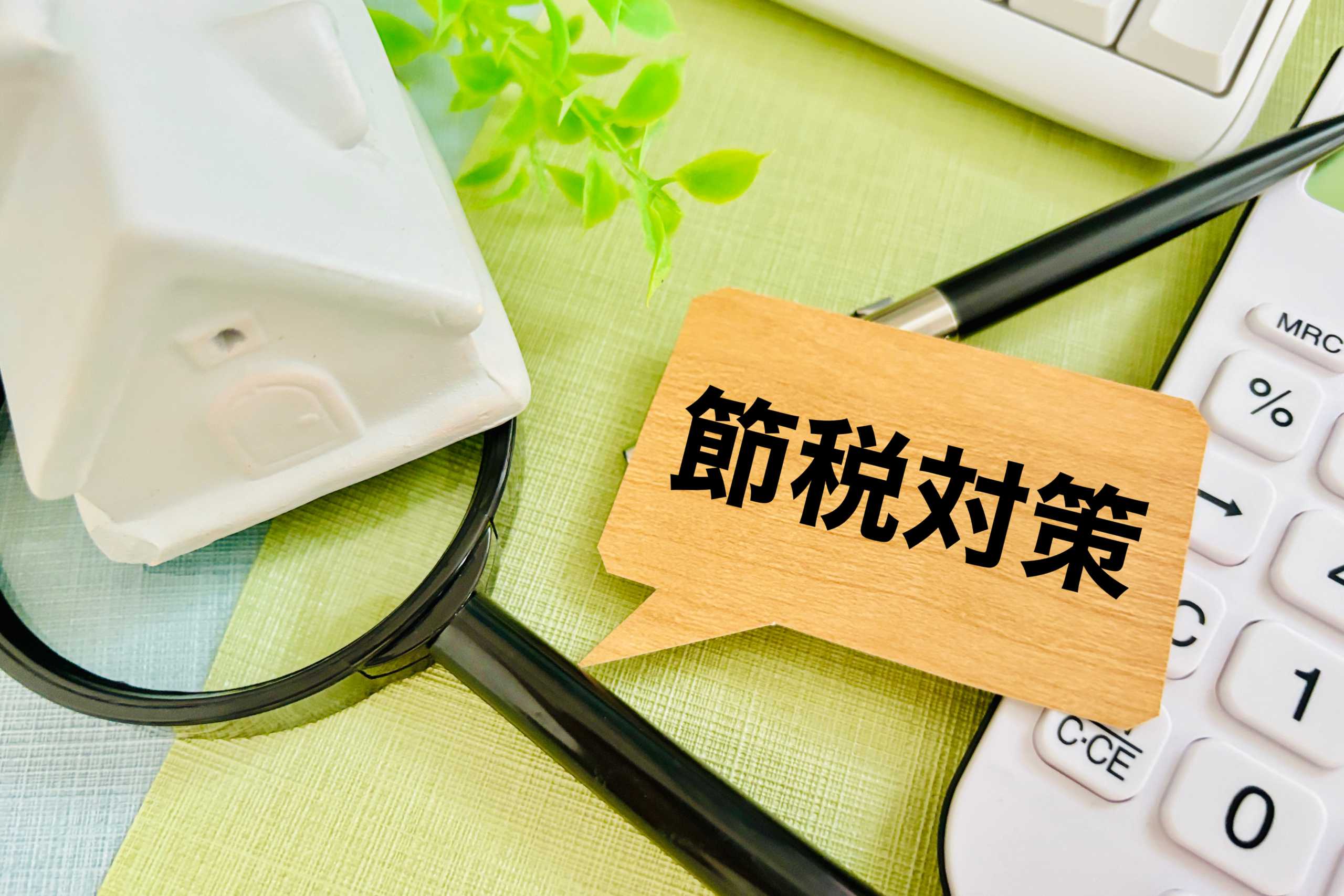 「節税対策」と書かれた吹き出し型のカードを中心に、白い家のオブジェ、虫眼鏡、電卓、ペンが並ぶデスクの上