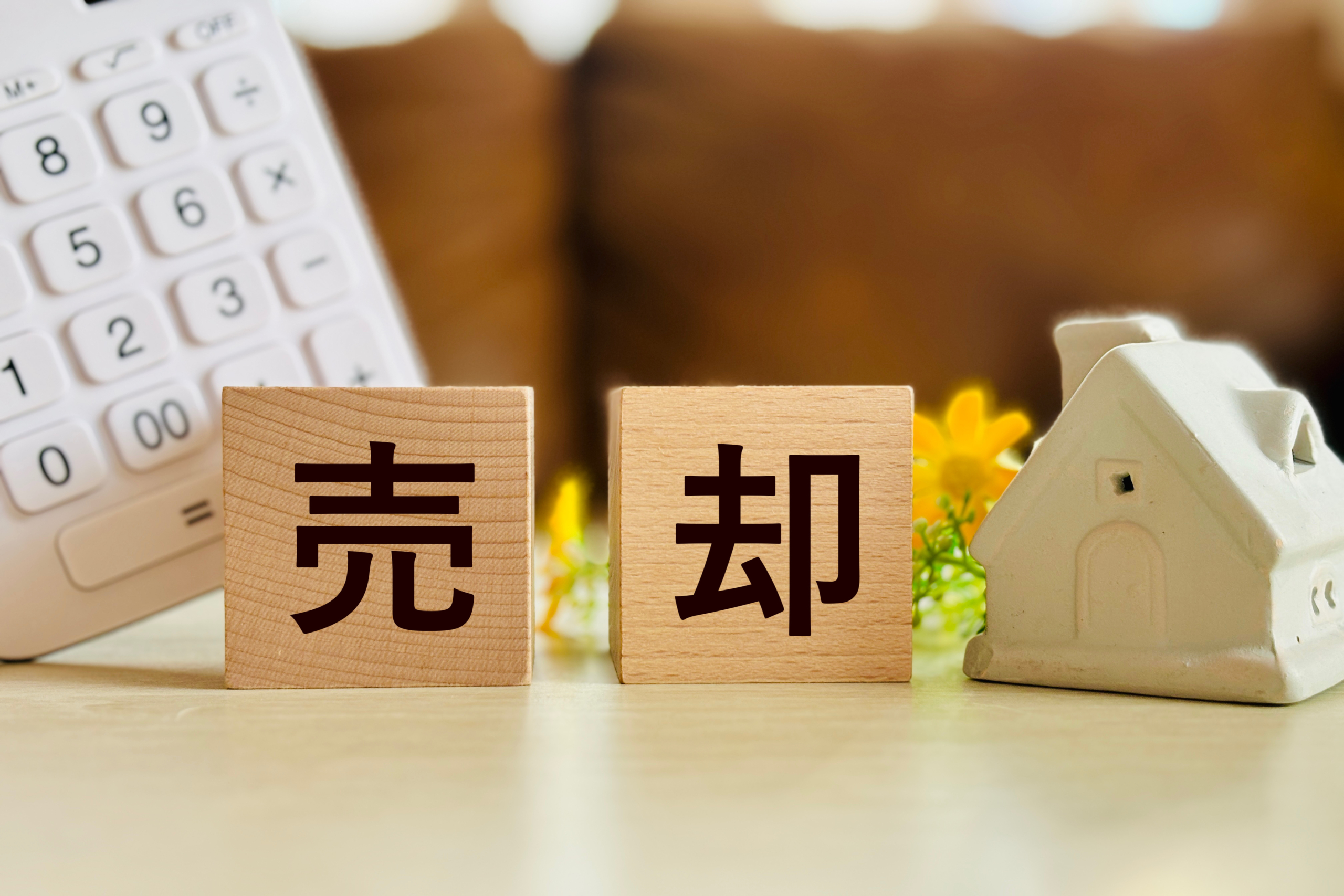 「売」「却」と書かれた木製ブロックを中心に、左側に白い電卓、右側に白い家のオブジェが置かれたテーブルの上